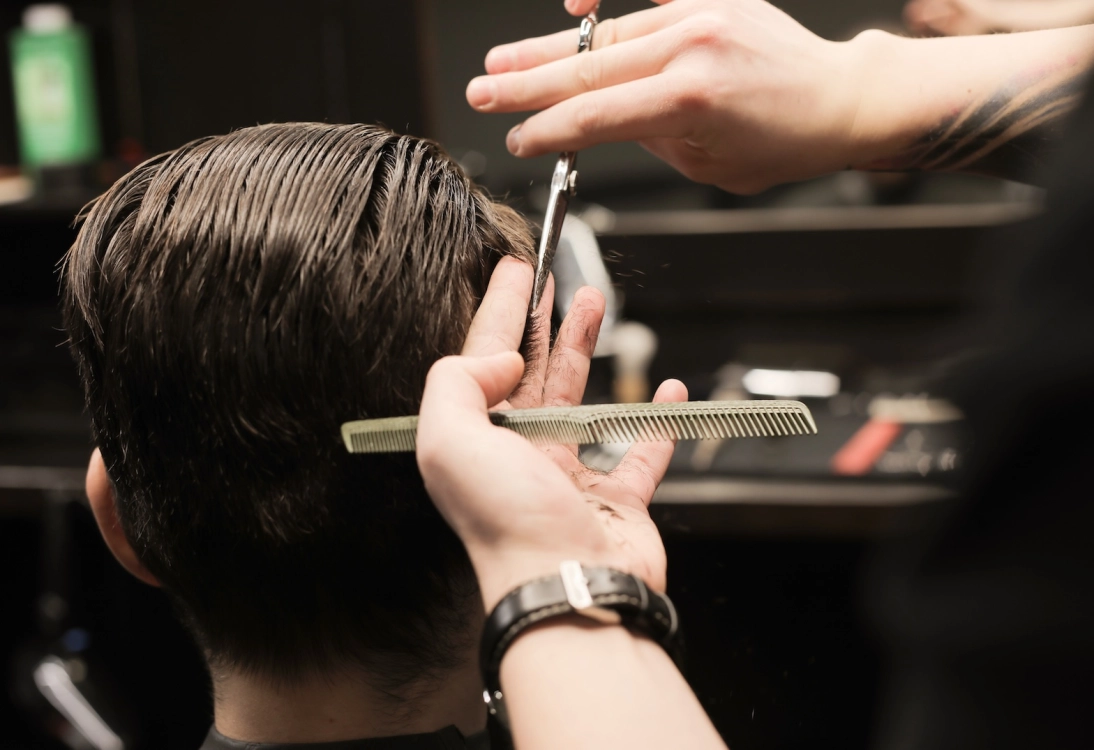 Neat movements. Close up photo of barber’s hands trimming hair of his client using scissors and comb.