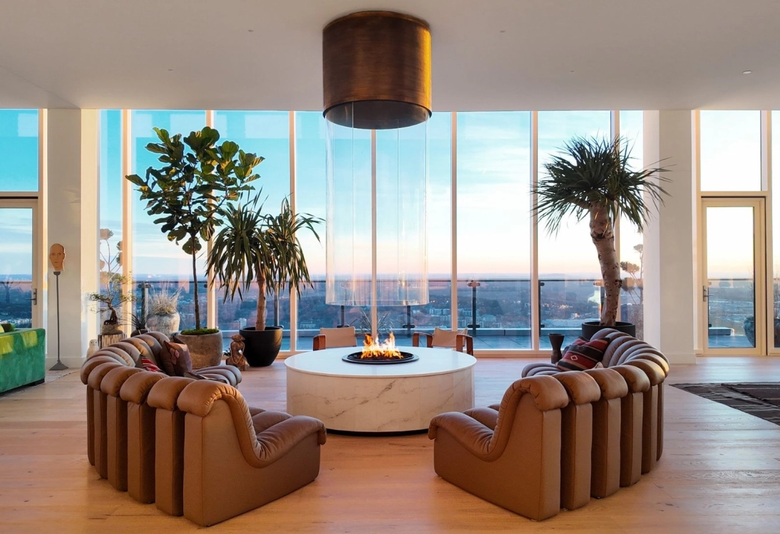 Modern living room with large curved brown sofas around a central fire pit and large windows showing cityscape.