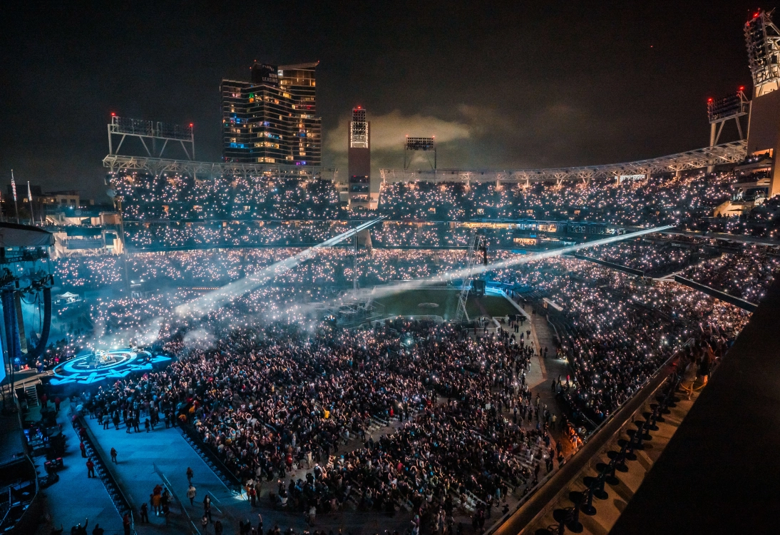 Nighttime concert in a large stadium with a crowd illuminated by phone lights and stage spotlights.