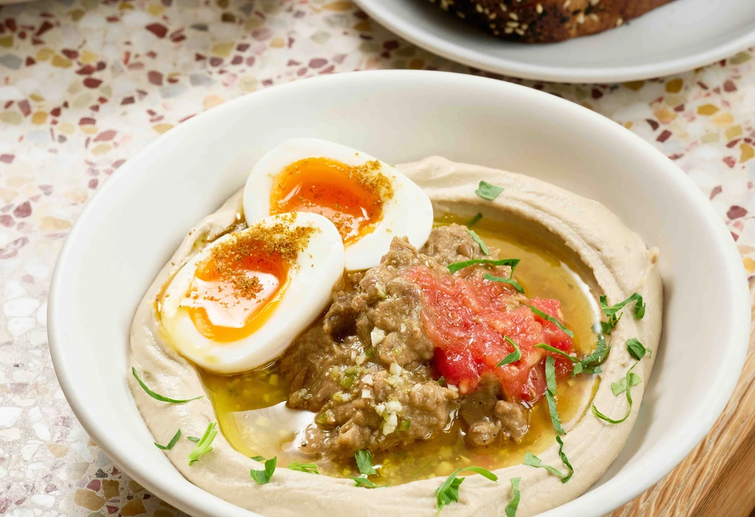 Bowl of hummus topped with soft-boiled egg, tomato, herbs, and a side of toasted bread and salad.