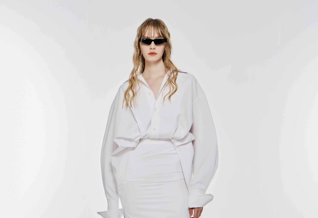 Woman wearing oversized white shirt, white midi skirt, black knee-high boots, and black sunglasses against white background.