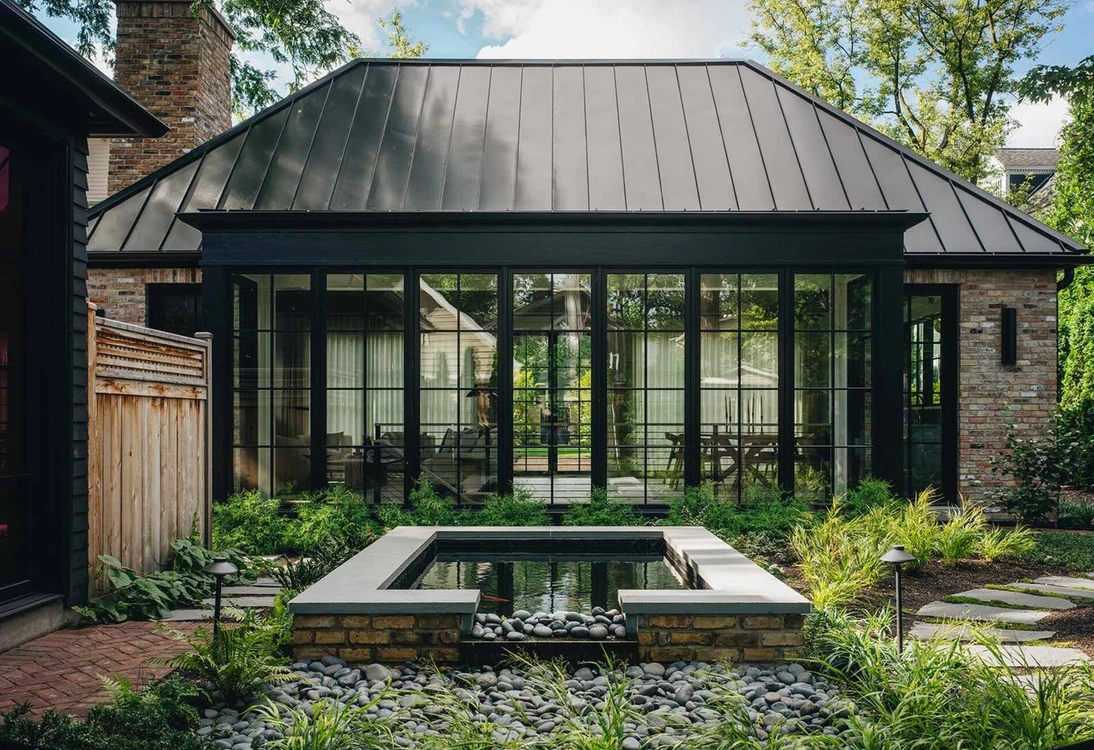 Modern garden patio with pond, surrounded by plants and a glass-walled building with a metal roof.