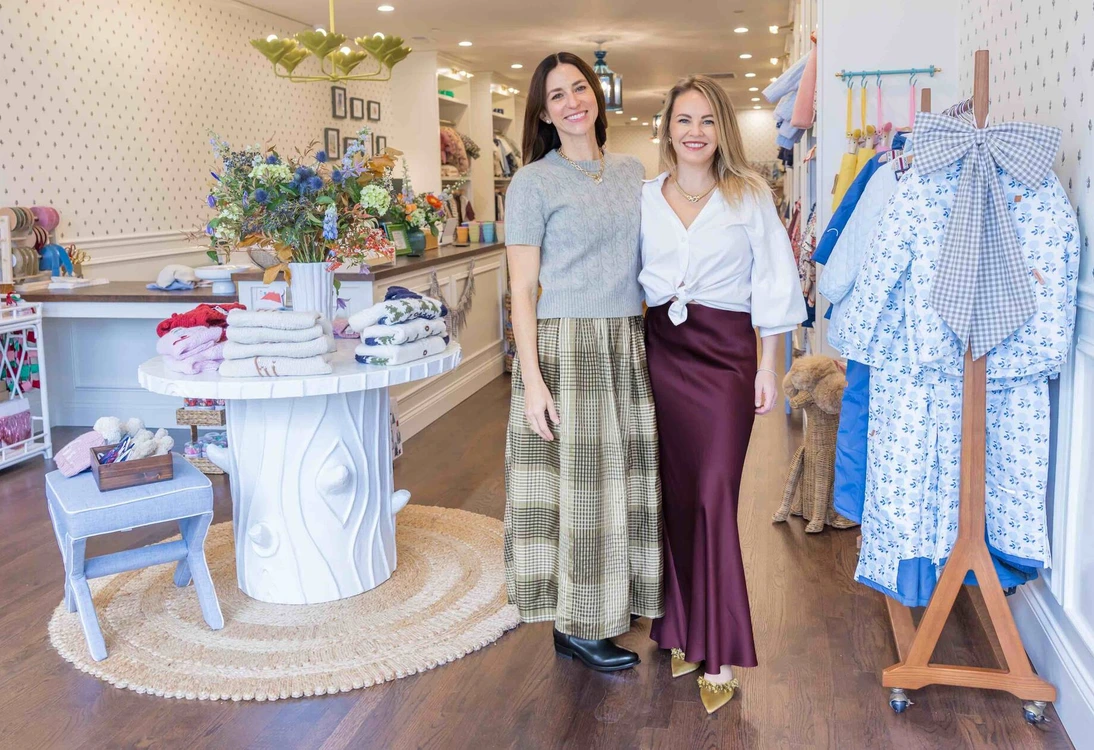 Two smiling women standing in a boutique clothing store with displayed clothes and decor.