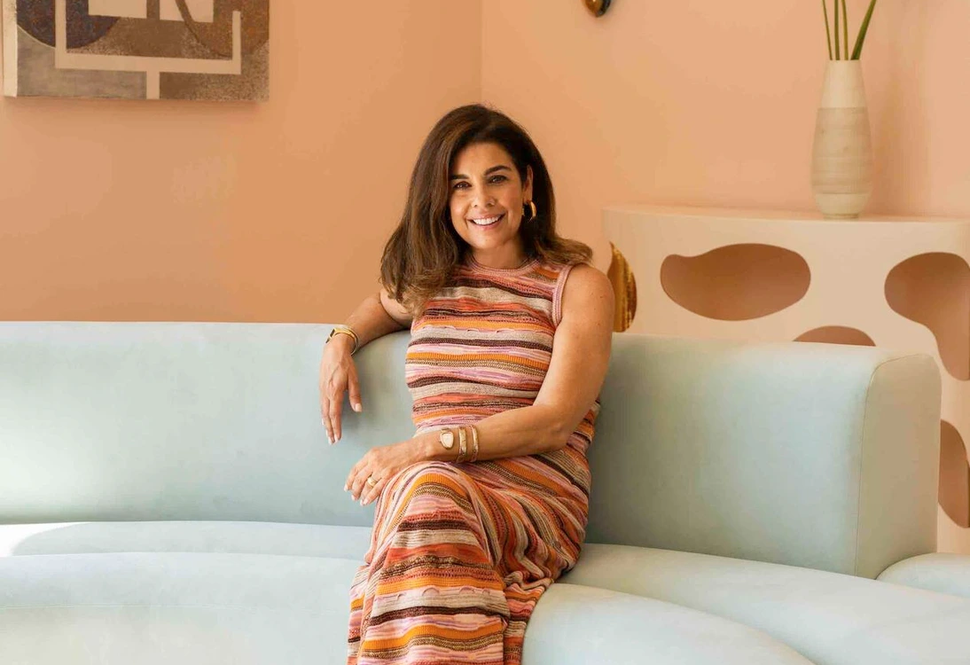 Woman smiling seated on curved light blue sofa in modern room with abstract art and vase of flowers.