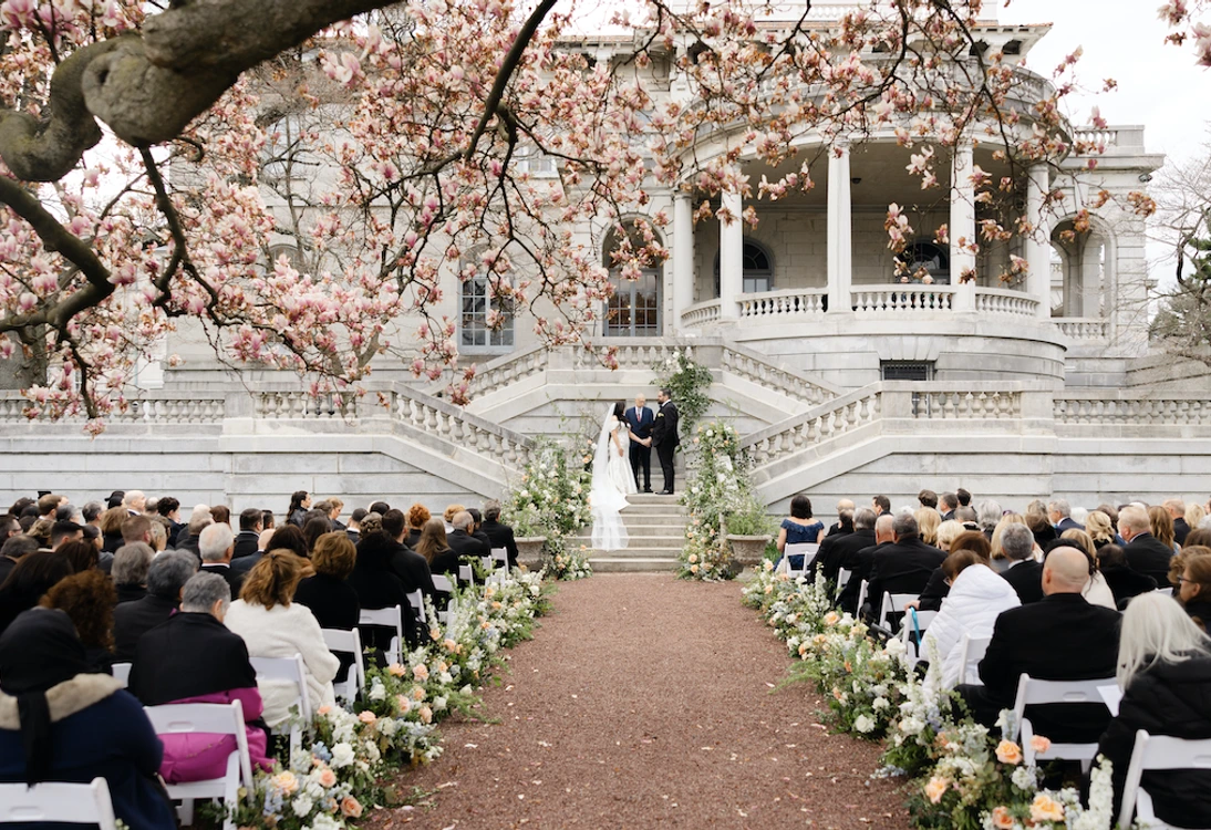 Outdoor wedding ceremony with bride, groom, officiant, guests seated, floral aisle decorations, and blooming tree branches.