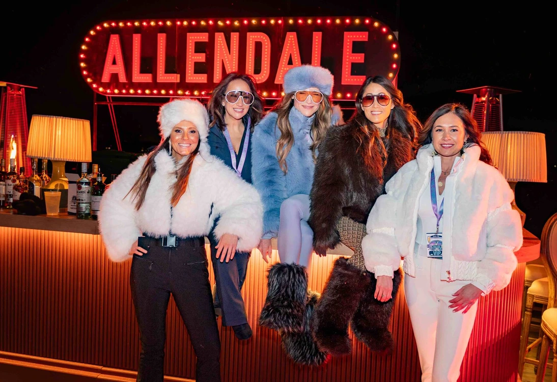Five women wearing winter outfits posing at a bar with a bright ALLENDALE sign in the background.