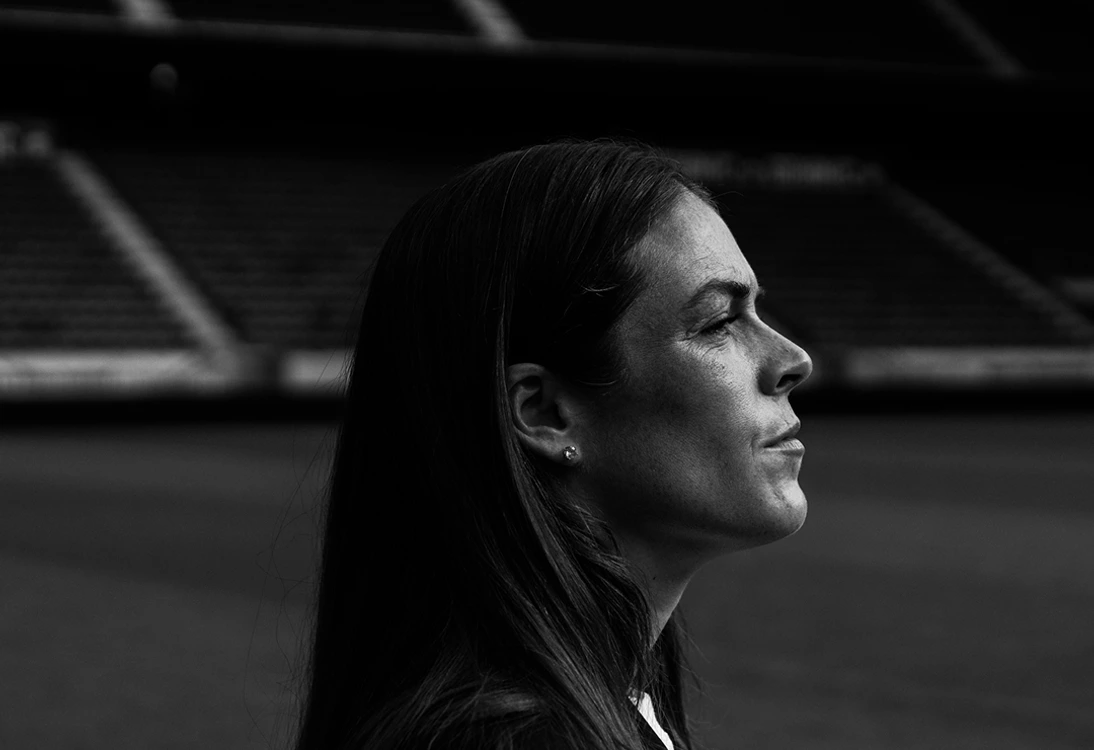 Black-and-white profile portrait of Kelley O’Hara on a soccer field, gazing forward with stadium stands blurred in the background.