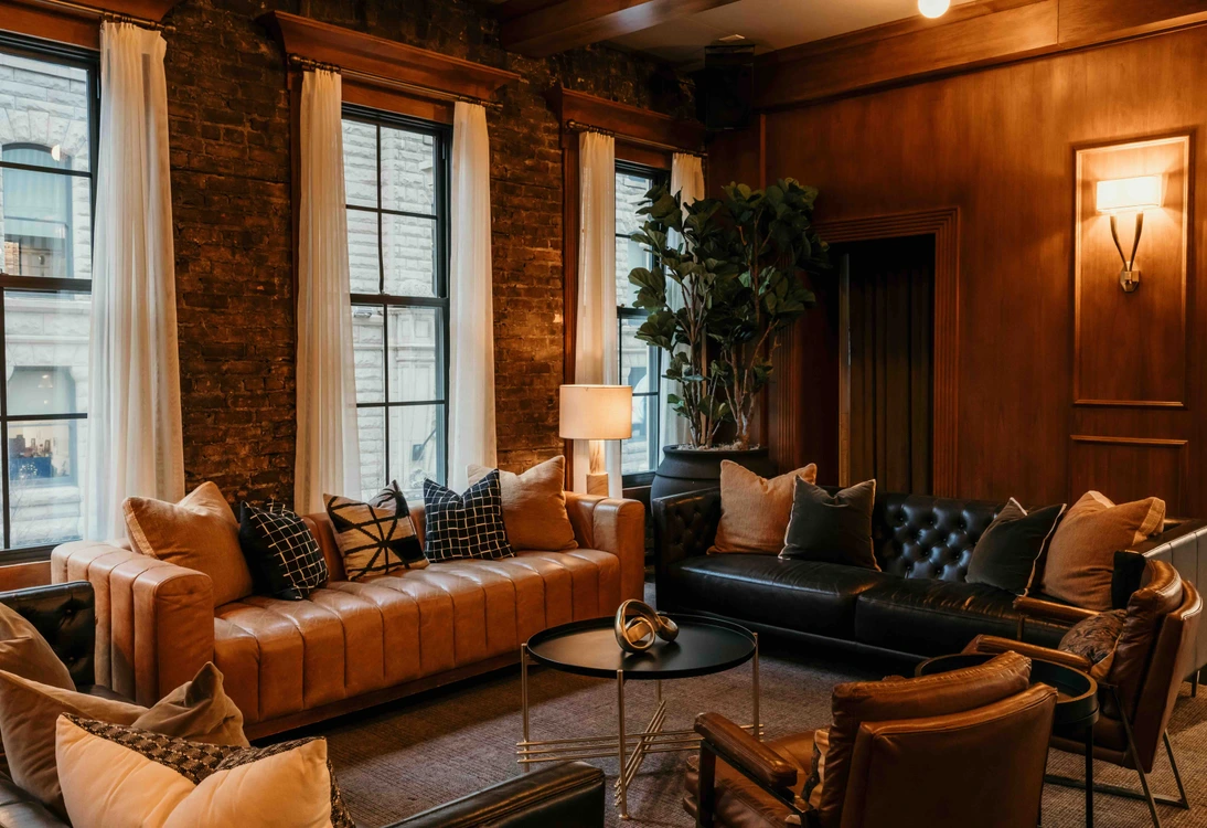 Cozy living room with leather sofas, pillows, large windows, wooden walls, and a round black coffee table.