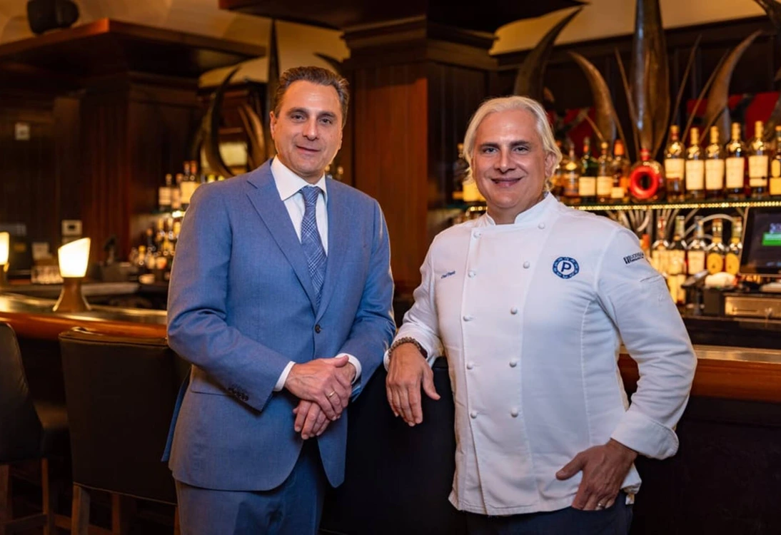 Two men, one in a blue suit and the other in a white chef's jacket, standing in a bar setting.