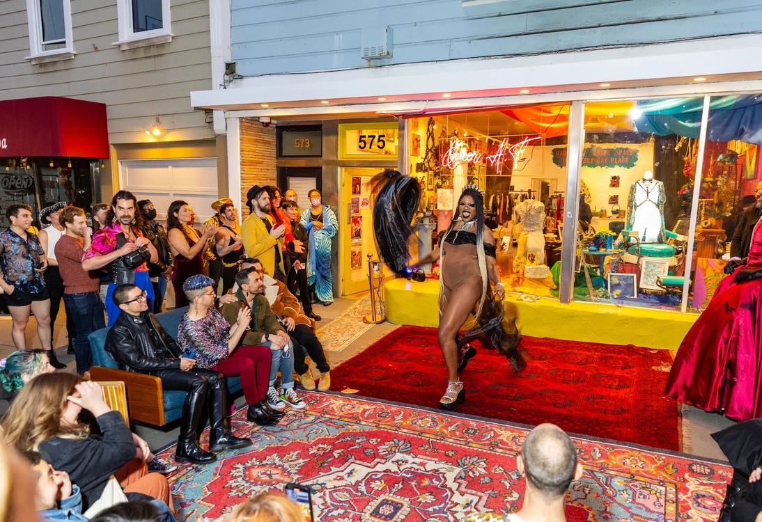 Drag performer walking on outdoor runway with seated and standing audience in front of colorful storefront.