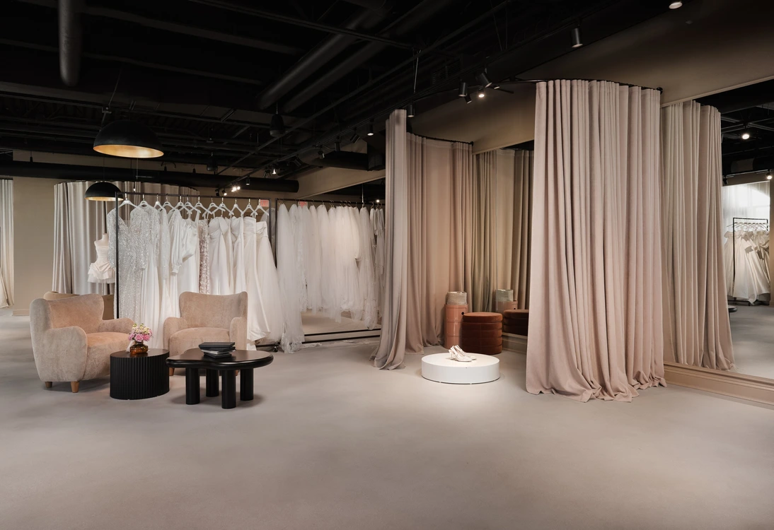 Interior of a bridal boutique with wedding dresses, fitting rooms with curtains, and seating area with chairs and tables.