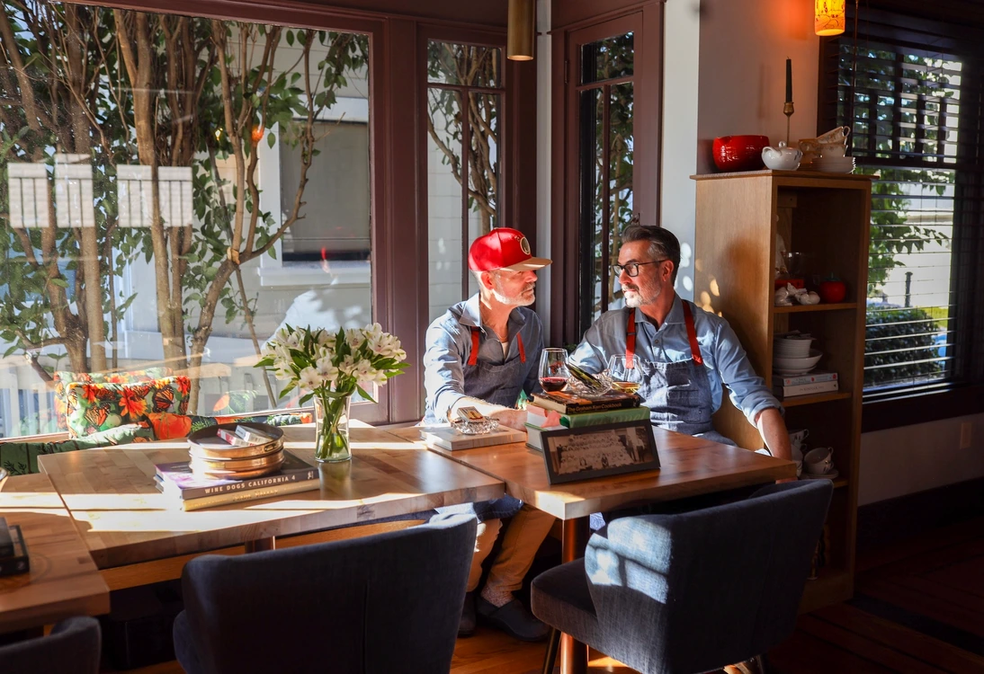 Two men sitting at a sunlit table in a cozy room, engaged in conversation.