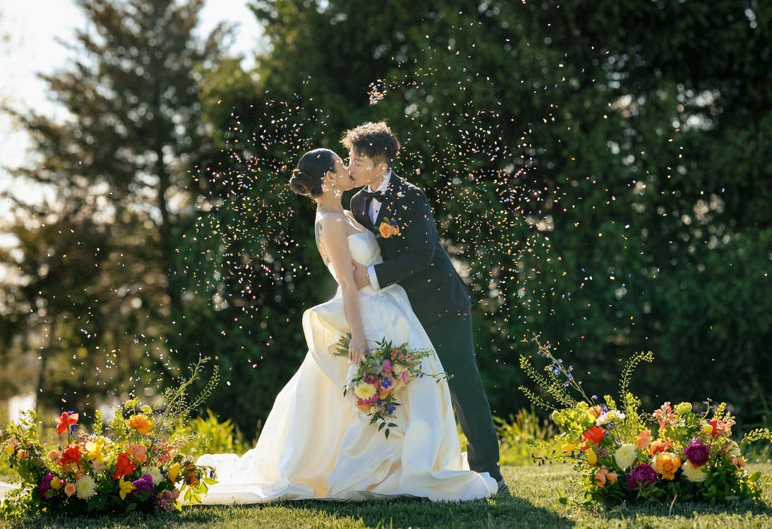 Bride and groom kissing outdoors during wedding ceremony with flower petals falling around them.