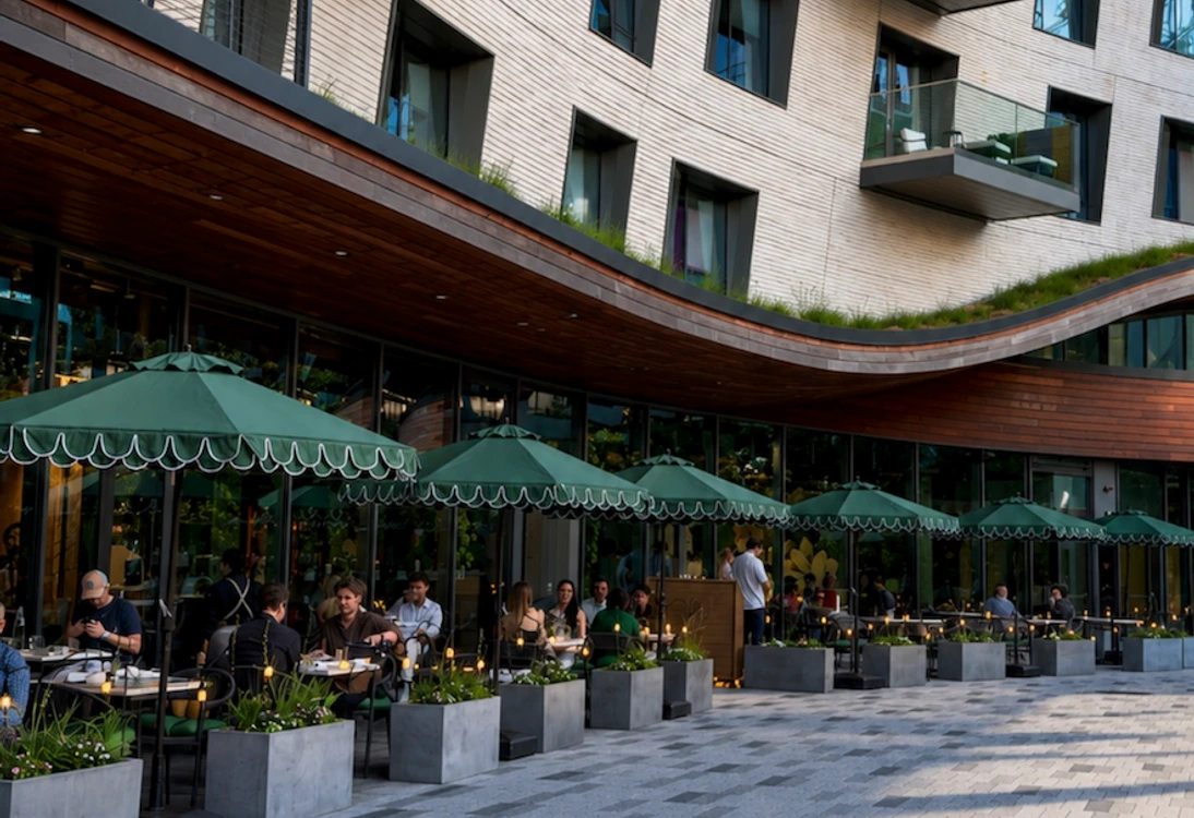 Outdoor cafe seating with green umbrellas beside a modern building with balconies and large windows.