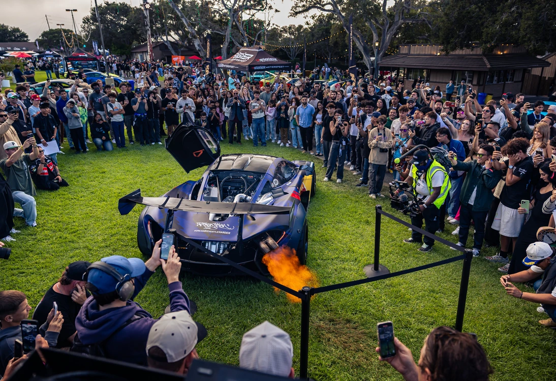 Crowd of people surrounding a sports car with flames coming from its exhaust at an outdoor event.