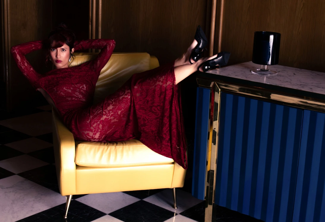 Woman in a red lace dress reclining on a yellow chair with feet on a blue and gold table.