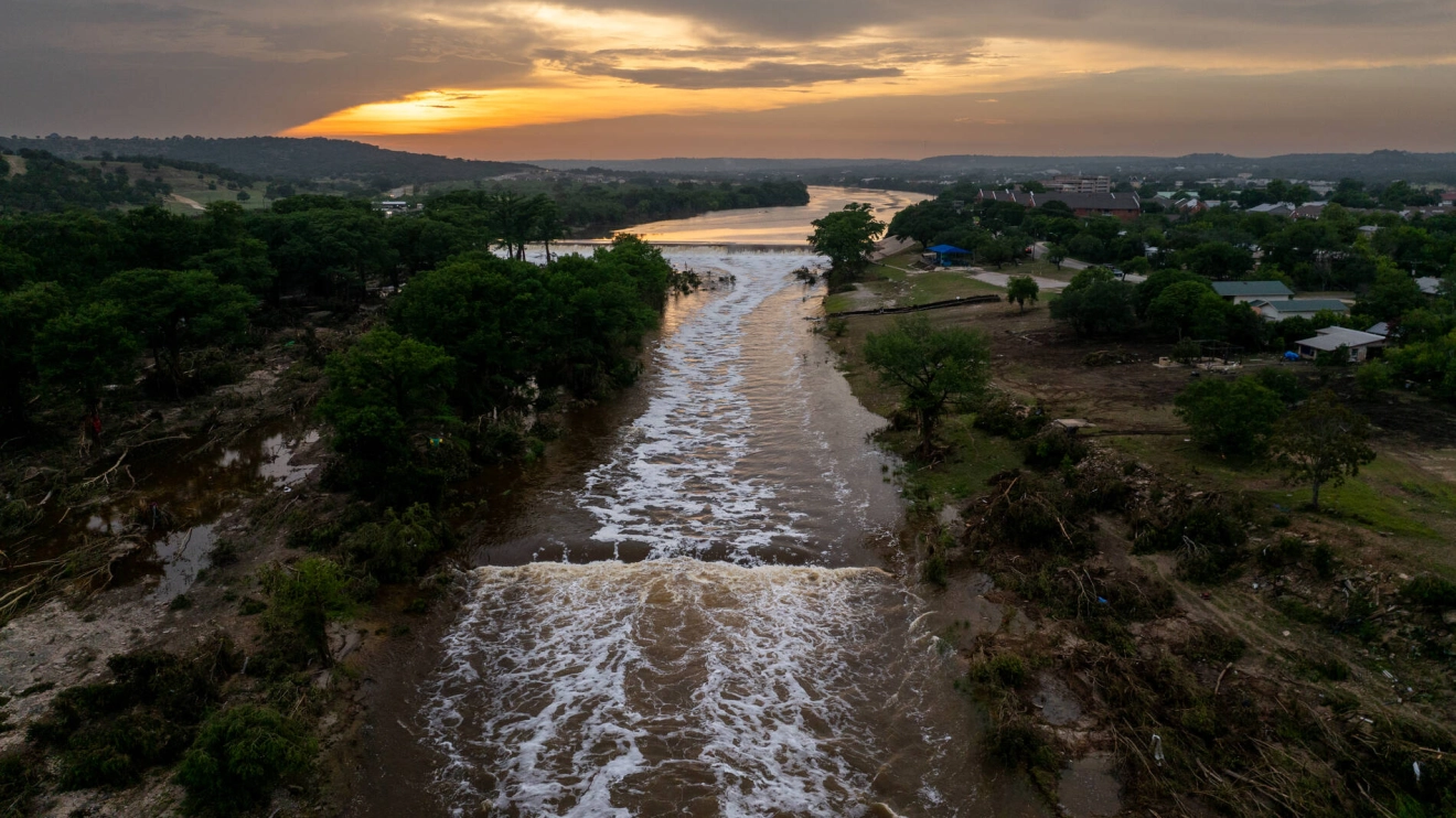 Aerial view of a river with flowing water at sunset, surrounded by greenery and a small settlement.