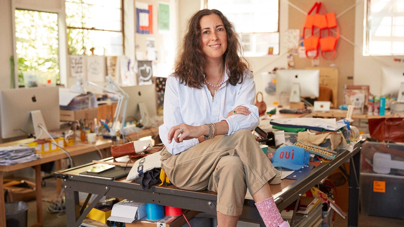Woman sitting on a cluttered worktable in a bright, casual office or studio space.