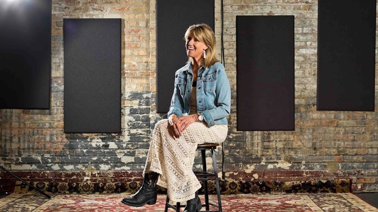 Woman sitting on a stool in a rustic room with brick walls, black panels, and a patterned carpet.