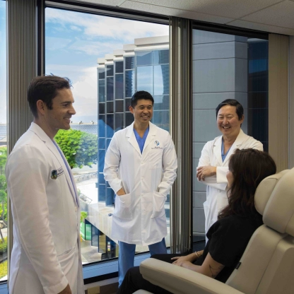 Three doctors in white coats converse with a patient in a medical office with large windows.