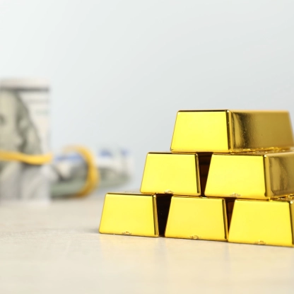 Stack of gold bars with blurred rolls of US dollar bills in the background on a flat surface.