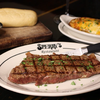 Grilled steak with herbs on a plate from Sperry's Restaurant, served with bread, butter, and loaded baked potato.