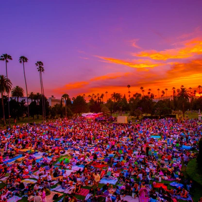 Large crowd gathered on a lawn at sunset with palm trees and colorful sky in the background.