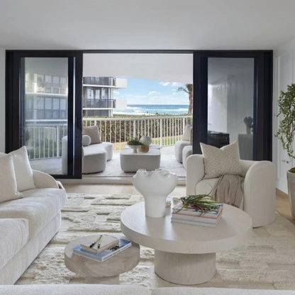 Modern, minimalist living room with cream-colored furniture and large sliding glass doors opening to a balcony with ocean view.