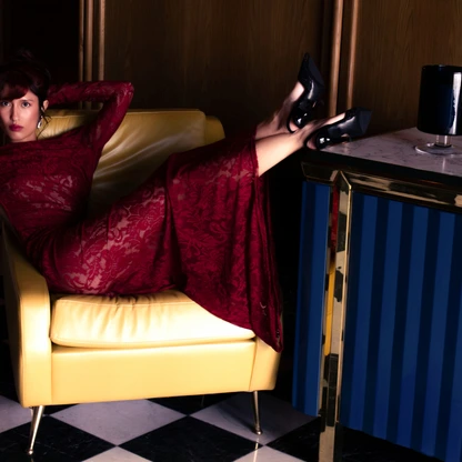 Woman in a red lace dress reclining on a yellow chair with feet on a blue and gold table.