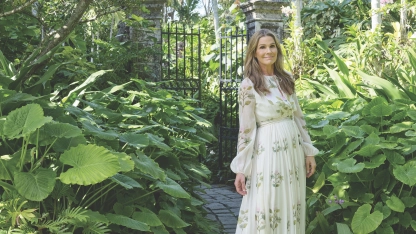 A woman in a floral dress stands amidst lush greenery near a garden gate.