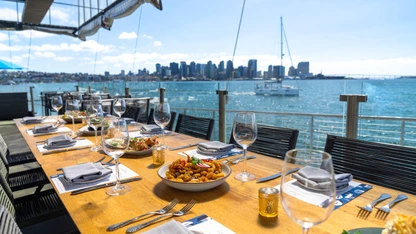Outdoor dining table set with food and wine glasses overlooking a city skyline and water with boats.