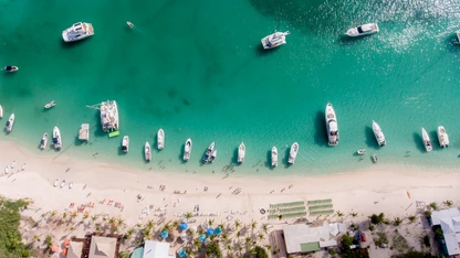 Aerial view of boats anchored near a sandy beach with people and tropical greenery.
