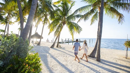 Couple walking hand-in-hand on a sunny tropical beach with palm trees and a pier in the background.