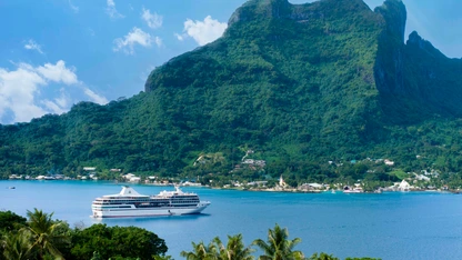 Cruise ship sailing near lush green mountain and tropical shoreline under blue sky.