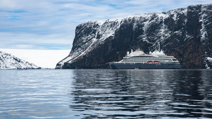 Cruise ship sailing near steep, snow-covered cliffs with calm water and cloudy sky.