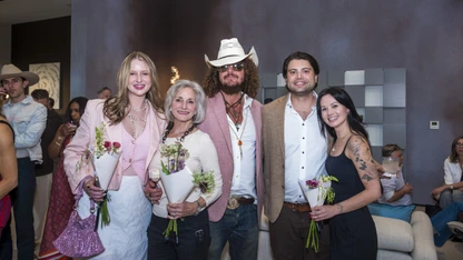 Group of five people posing indoors, three women holding flowers, one man wearing a cowboy hat and sunglasses.