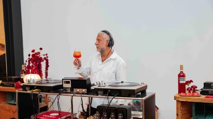 DJ in white shirt holding a cocktail glass, standing behind turntables and audio equipment.