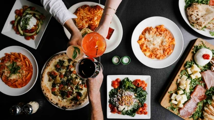 Top view of a table with Italian dishes including pizza, pasta, salad, and drinks being toasted by hands.