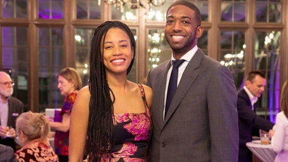 A smiling couple dressed formally at an indoor event with guests in the background.