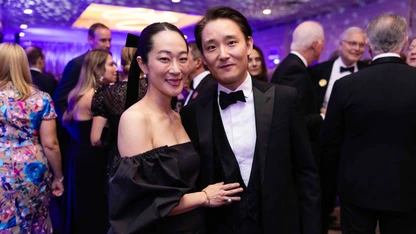 Couple dressed formally, man in tuxedo, woman in black off-shoulder dress, at elegant indoor event with guests.