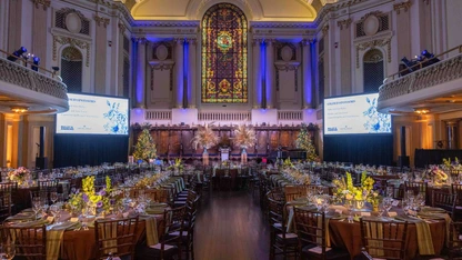 Elegant banquet hall with decorated tables, stained glass window, and large screens for an event.