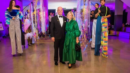 Couple posing at event with performers on stilts holding colorful decorations in brightly lit space.