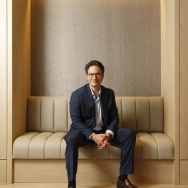 A man in a dark suit sits on a bench in a well-lit, modern interior.