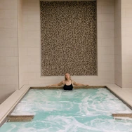 Woman relaxing in an indoor pool with tiled wall and beige stone surroundings.