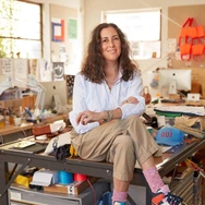 Woman sitting on a cluttered worktable in a bright, casual office or studio space.