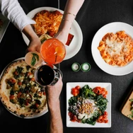 Top view of a table with Italian dishes including pizza, pasta, salad, and drinks being toasted by hands.