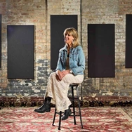 Woman sitting on a stool in a rustic room with brick walls, black panels, and a patterned carpet.
