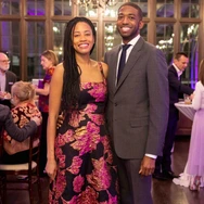 A smiling couple dressed formally at an indoor event with guests in the background.
