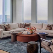 Modern living room with large beige sectional sofa, round coffee table with flowers, and panoramic city view windows.