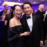 Couple dressed formally, man in tuxedo, woman in black off-shoulder dress, at elegant indoor event with guests.